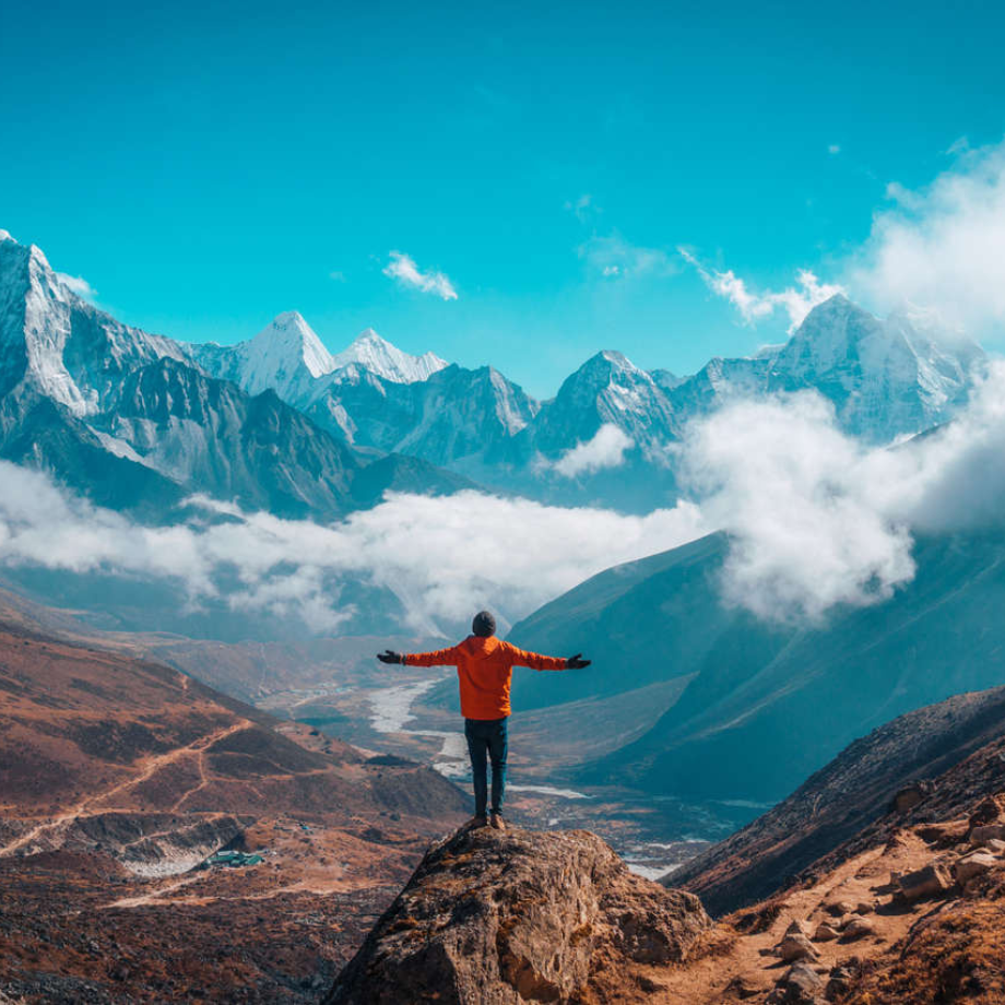 A person in an orange jacket stands on a rocky cliff with arms outstretched, overlooking a vast mountain valley with snow-capped peaks under a bright blue sky and scattered clouds.