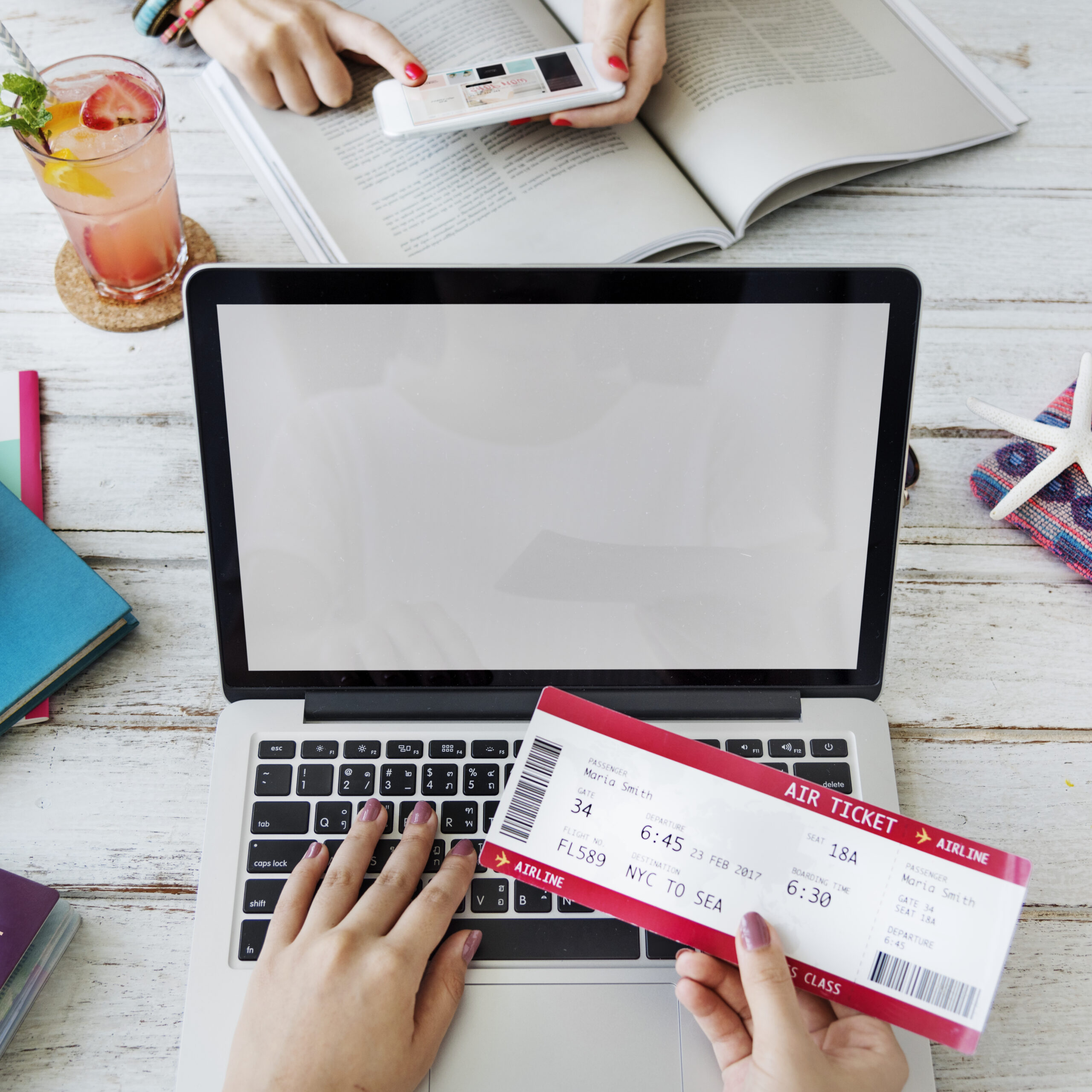 A person holds an airline ticket while using a laptop on a white wooden table with notebooks, a pen, a drink, and another person reading a magazine in the background.